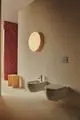 Badezimmer mit Wand-Bidet und wandhängendem WC von Ideal Standard. Eine runde Leuchten bringt sanftes Licht in den modernen Raum mit beige und roten Akzenten.